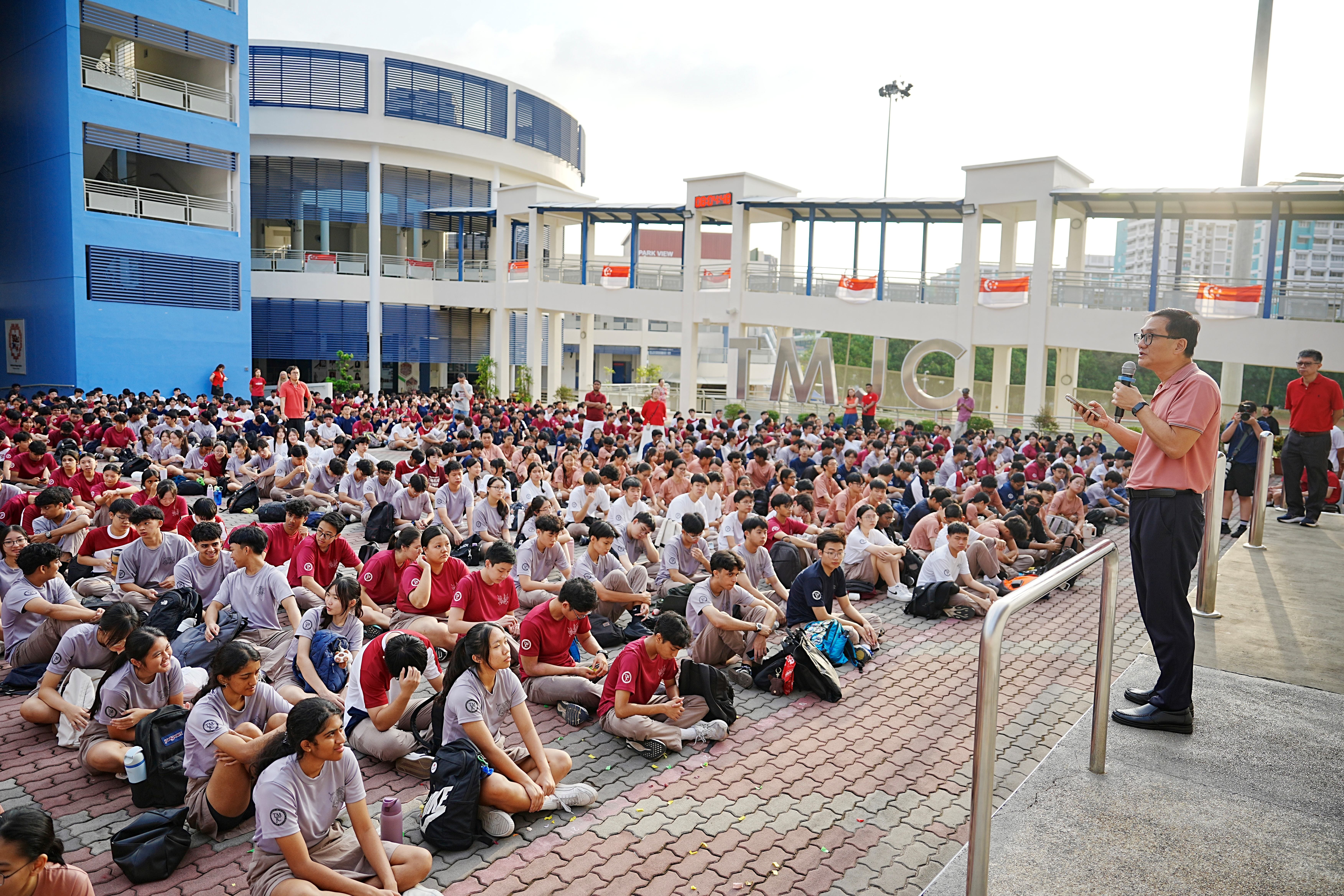The principal addresses a large seated crowd of students outdoors in a school courtyard, with the "TMJC" lettering visible on the building behind him.